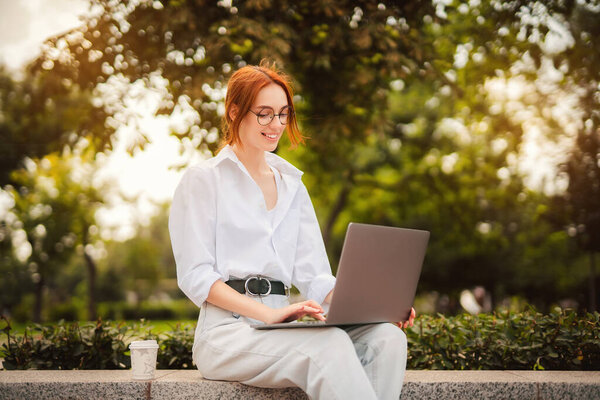 Beautiful redhead young woman sitting in the park and using laptop. Student. University. Freelance. Wearing in glasses casual jeans and white shirt. Coffee to go. Paper cup. Looking in laptop. Smiling