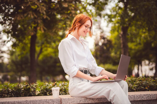 Beautiful redhead young woman sitting in the park and using laptop. Student. University. Freelance. Wearing in glasses jeans and white shirt. Coffee to go. Paper cup. Looking in laptop. Smiling 