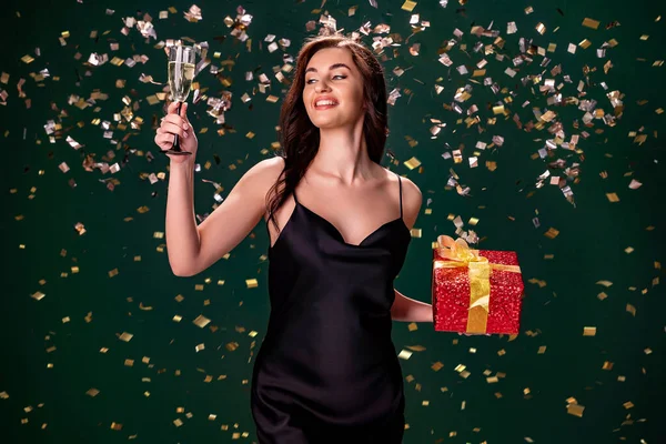 Aufgeregt hält eine Frau im schwarzen Kleid die rote Geschenkbox und ein Glas Champagner in der Hand. Partyzeit. Urlaubskonzept — Stockfoto