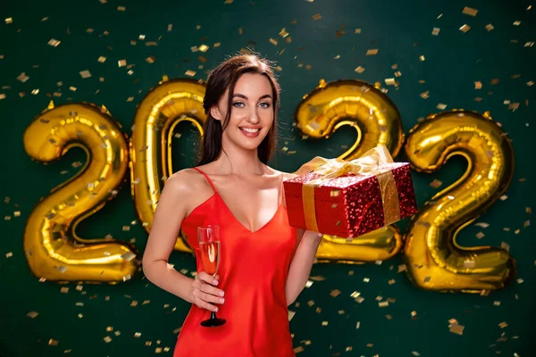 Aufgeregt hält eine Frau im roten Kleid die rote Geschenkschachtel und ein Glas Champagner in der Hand. Partyzeit. Urlaubskonzept — Stockfoto