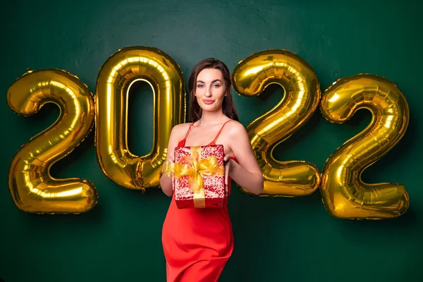 Улыбающаяся брюнетка в красном платье поднимает подарочную красную коробку. Золотые воздушные шары NEW YEAR 2022. — стоковое фото