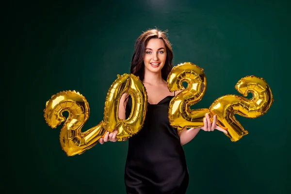 Jovem mulher em vestido de cocktail com maquiagem brilhante celebrando Ano Novo 2022 e segurando balões dourados 2022 em mãos sobre fundo verde — Fotografia de Stock