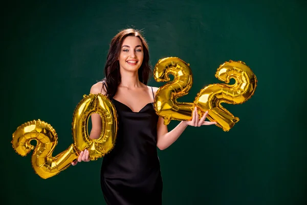 Junge Frau im Cocktailkleid mit hellem Make-up feiert Neujahr 2022 und hält goldene Luftballons in den Händen auf grünem Hintergrund — Stockfoto