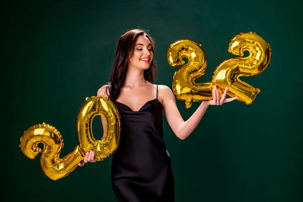 Mujer joven en vestido de cóctel con maquillaje brillante que celebra el Año Nuevo 2022 y la celebración de globos de oro 2022 en las manos sobre fondo verde — Foto de Stock