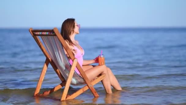 Mujer hermosa y sexy en traje de baño rosa sentada en una silla de sol en la playa — Vídeos de Stock