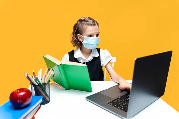 Seriös skolflicka i mask gör sina läxor med bok och laptop. Distansutbildning — Stockfoto