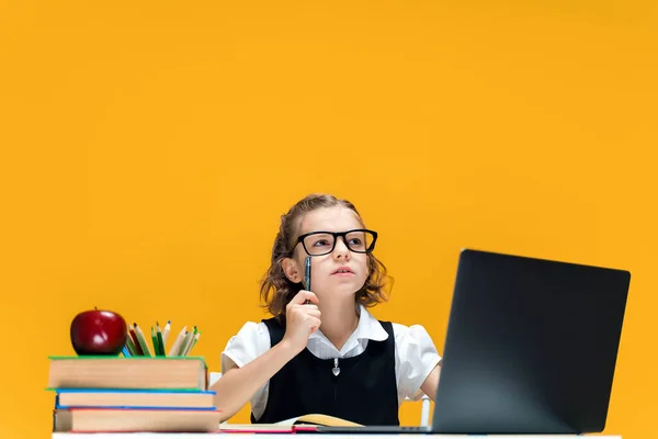 Écolière sérieuse en lunettes levant le stylo. Assis au bureau avec un ordinateur portable. Enseignement à distance — Photo