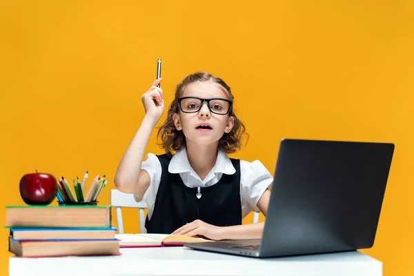 Vážná školačka v brýlích zvedá pero. Sedí za stolem s laptopem. Vzdělání na dálku — Stock fotografie