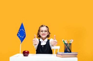 Masada oturan mutlu beyaz kız öğrenci başparmağıyla işaret ediyor. İngilizce dersi. AB bayrağı