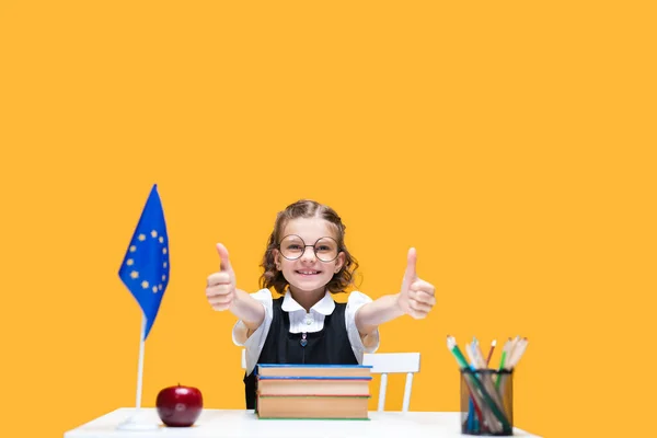 Happy caucasian schoolgirl sitting at the desk gesturing thumbs up. English lesson. EU flag