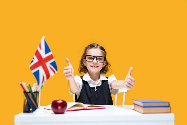 Happy caucasian schoolgirl sitting at the desk showing thumbs up. English lesson Great Britain flag — Stock Photo, Image