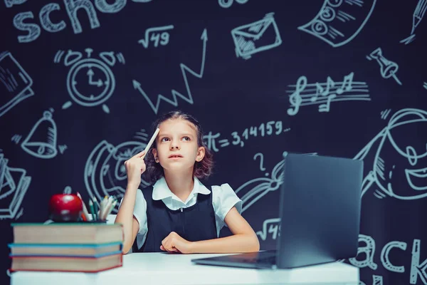 Серйозна вдумлива школярка піднімає ручку. Сидячи за столом з ноутбуком. Дистанційне навчання — стокове фото