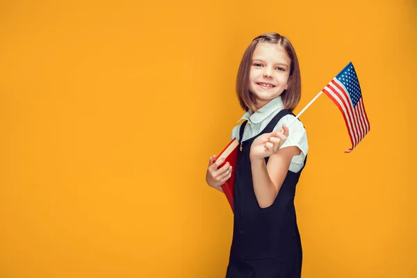 Leende kaukasiska skolflicka hålla amerikansk flagga och boka i händerna på gul bakgrund. Förenta staternas flagga — Stockfoto