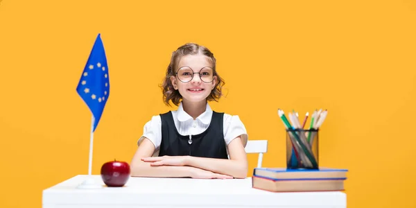 Smiling happy caucasian schoolgirl sitting at the desk during English lesson. European Union flag