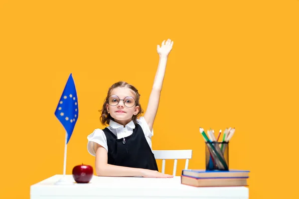 Caucasian schoolgirl raising hand up sitting at the desk during lesson. English lesson. Europe flag — Stock Photo, Image