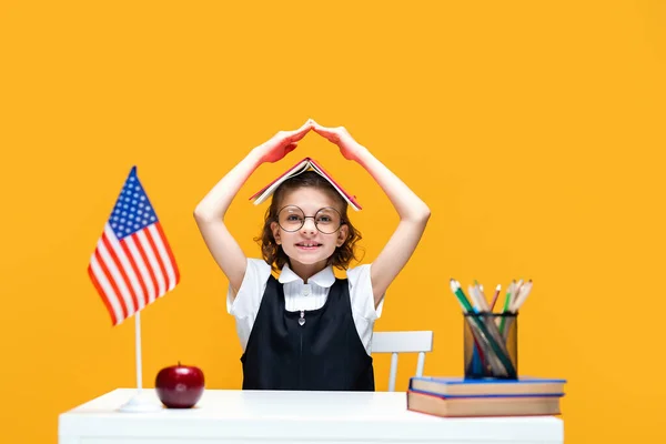 Schoolgirl in fun sitting at the desk holding book on her head. English lesson. USA flag