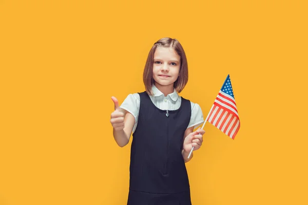 Malá roztomilá školačka s americkou vlajkou ukazuje palec nahoru na žlutém pozadí. Jako Amerika. — Stock fotografie