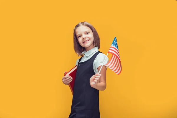 Roztomilé usmívající se běloška školačka drží americkou vlajku a knihu na žlutém pozadí. Vlajka USA — Stock fotografie
