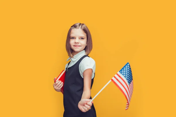 Roztomilé běloška školačka ukazuje americkou vlajku a drží knihu na žlutém pozadí. Vlajka USA — Stock fotografie