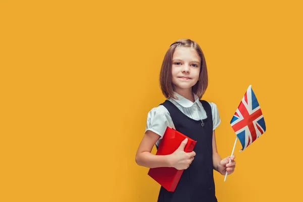 Little cute schoolgirl holding United Kingdom flag and book studying English on yellow background.