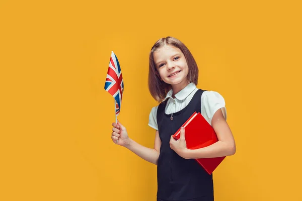 Little schoolgirl holding United Kingdom flag and book studying English on yellow background.