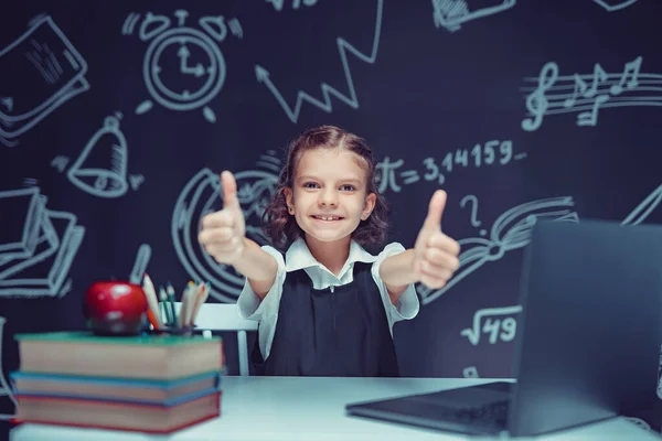 Emocionální školačka ukazující palce nahoru, nacházející inspiraci nebo řešení, sedící za stolem s laptopem a provádějící domácí úkoly — Stock fotografie
