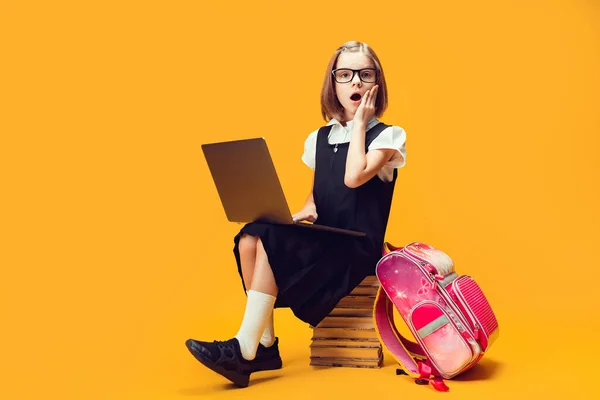 Full length shocked schoolgirl sit on the stack of books with PC Κοιτάξτε την κάμερα. Εκπαίδευση των παιδιών — Φωτογραφία Αρχείου