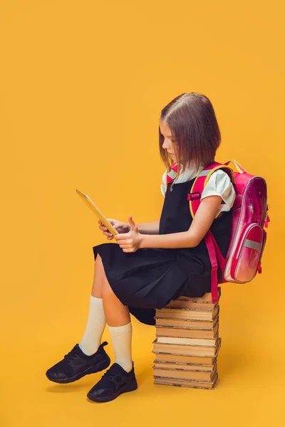 Full length schoolmeisje in uniform zitten op de stapel boeken werk op tablet. Onderwijsconcept voor kinderen. — Stockfoto