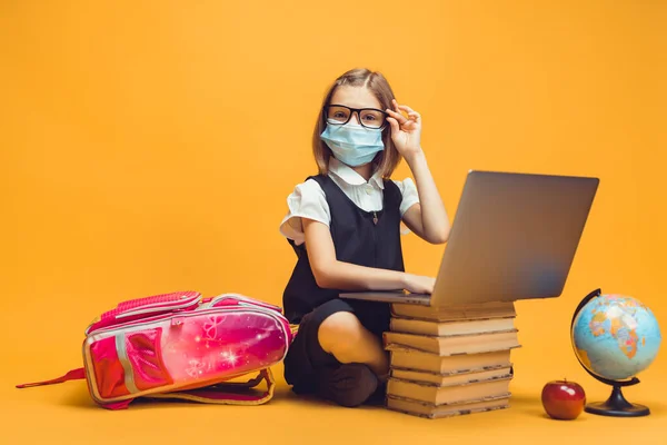 Skolflicka i medicinsk mask sitter bakom en bunt böcker och laptop. Utbildning för barn vid pandemi — Stockfoto