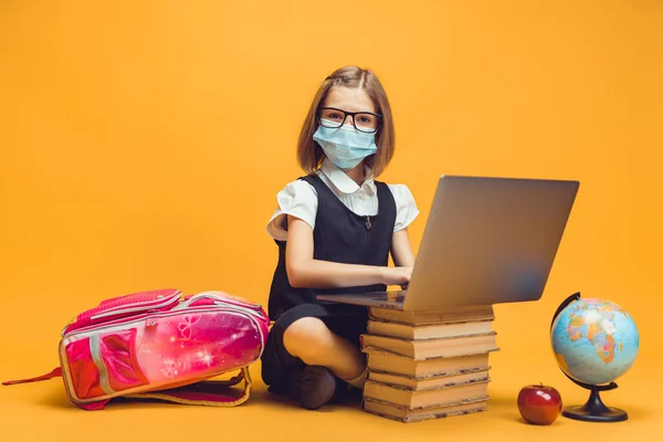 Skolflicka i medicinsk mask sitter bakom en bunt böcker fungerar på laptop. Utbildning för barn vid pandemi — Stockfoto