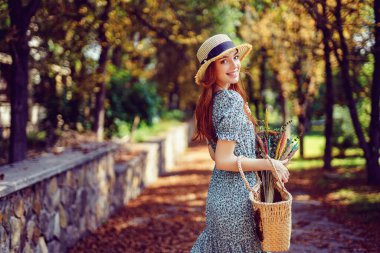 Güneşli bir günde gülen kızıl saçlı kız, elinde el çantası tutarken sonbahar parkında tek başına yürüyor. 