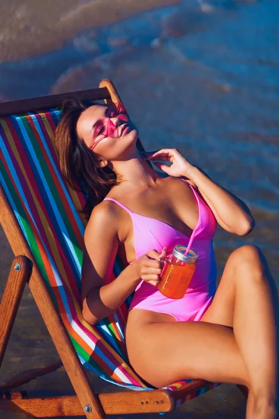 Привлекательная рыжая девушка на шезлонге загорает у моря со стаканом сока в руке — стоковое фото