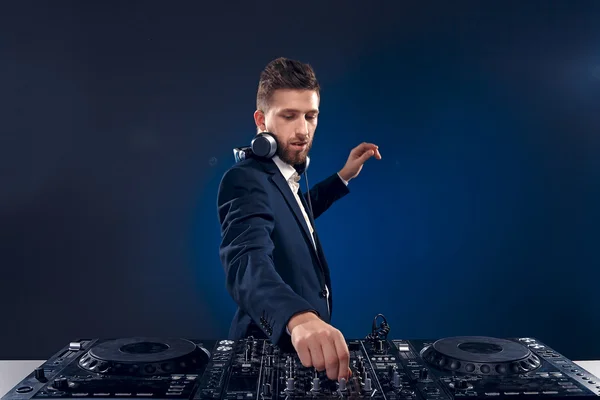 Nahaufnahme Porträt eines selbstbewussten DJs mit stylischer Frisur und Kopfhörern im Nacken, der Musik am Mischpult mixt, während er isoliert auf dunkelblauem, cyanischem Hintergrund steht — Stockfoto