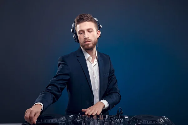 Primer plano retrato de DJ confiado con estilo de pelo elegante y auriculares en el cuello mezclando música en el mezclador, mientras que de pie aislado en azul de color oscuro, fondo cian —  Fotos de Stock