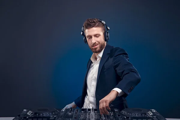 Closeup portrait of confident DJ with stylish hair style and headphones on neck mixing music on mixer while standing isolated on dark colored blue, cyan background