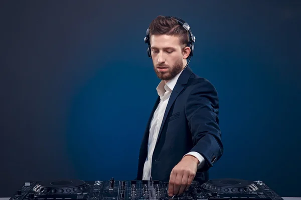 Retrato de close-up de DJ confiante com estilo de cabelo elegante e fones de ouvido no pescoço misturando música no mixer enquanto estava isolado em azul escuro, fundo ciano — Fotografia de Stock