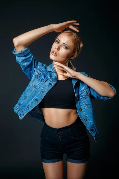 Attractive young blond girl wearing high waisted blue shorts and a black tank top, on a dark studio background.