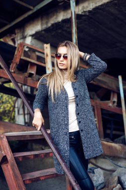 Bir güzel genç sarışın kadın gri ceket, güneş gözlüğü, portresi endüstriyel bir arka planda deri pantolon. Terk edilmiş metal fabrika.