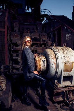 Bir güzel genç sarışın kadın gri ceket, güneş gözlüğü, portresi endüstriyel bir arka planda deri pantolon. Terk edilmiş metal fabrika.