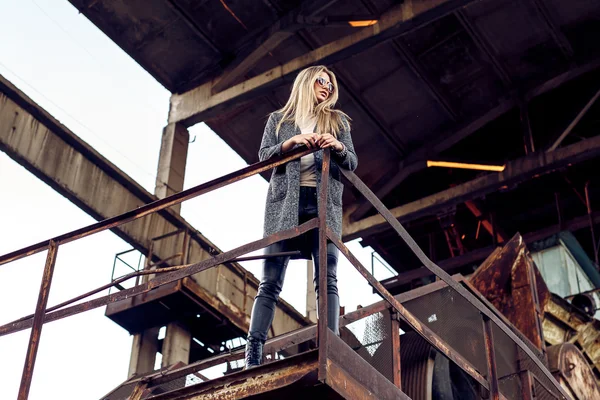 Retrato de uma bela jovem loira mulherem casaco cinza, óculos de sol, calças de couro em um fundo industrial. Fábrica de metal abandonada . — Fotografia de Stock