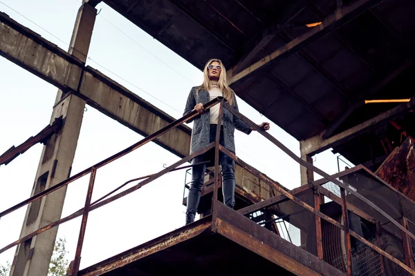 Retrato de uma bela jovem loira mulherem casaco cinza, óculos de sol, calças de couro em um fundo industrial. Fábrica de metal abandonada . — Fotografia de Stock