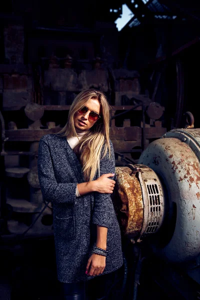 Retrato de uma bela jovem loira mulherem casaco cinza, óculos de sol, calças de couro em um fundo industrial. Fábrica de metal abandonada . — Fotografia de Stock