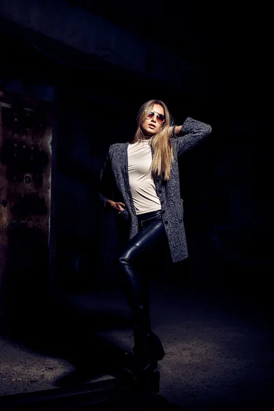 Portrait d'une belle jeune femme blonde en manteau gris, lunettes de soleil, pantalon en cuir dans un fond industriel. Usine de métal abandonnée . — Photo