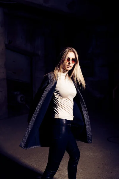 Portrait d'une belle jeune femme blonde en manteau gris, lunettes de soleil, pantalon en cuir dans un fond industriel. Usine de métal abandonnée . — Photo