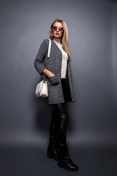 Sexy Beauty Girl. Fashion Blonde. Portrait of a girl dressed in grey coat and sunglasses, wearing a beige bag posing on a grey background. — Zdjęcie stockowe