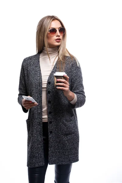 Chica joven delgada de moda en abrigo gris y gafas de sol, de pie charlando en un teléfono móvil mientras sostiene una taza de café para llevar en su otra mano, aislado en blanco — Foto de Stock