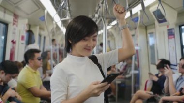 Çekici gülümseyen Asyalı kadının portresi tırabzanı tutuyor ve arkadaşlarıyla sohbet etmek veya toplu taşıma araçlarında cep telefonuyla gezinmek için akıllı telefon kullanıyor.
