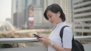 Kulaklıklı Asyalı bir kadının portresi müzik dinliyor ve sohbet için akıllı telefon kullanıyor ya da caddede geziniyor. Günlük hayatta ve seyahatte teknoloji.