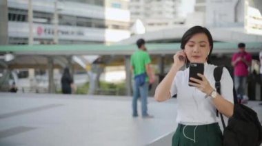 Kulaklıklı mutlu Asyalı kadın portresi müzik dinliyor, akıllı telefon dansı yapıyor ve caddede yürüyor. Günlük hayatta ve seyahatte teknoloji.