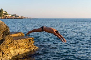 Kayalık bir sahilden suya atlayan kaslı bir sporcu. Arka planda deniz ufku